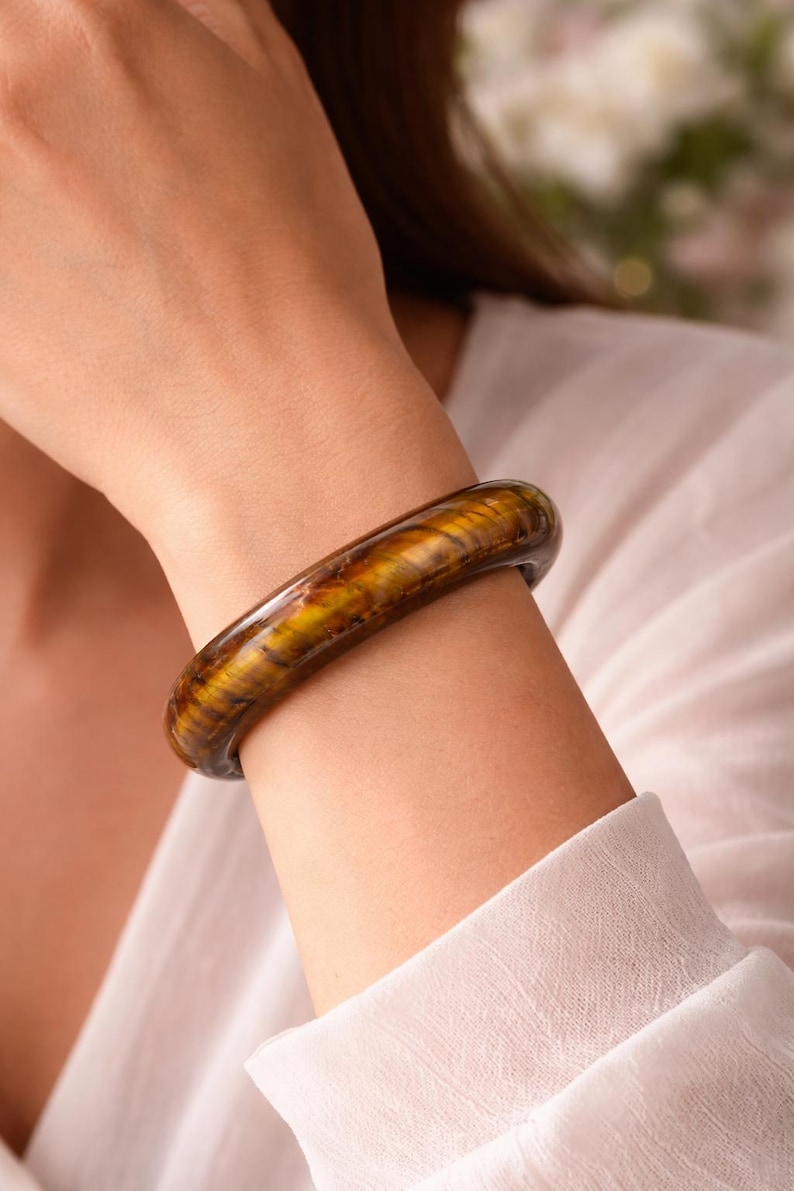 Puede incluir: Un brazalete r&iacute;gido pulido hecho de piedra ojo de tigre, con una superficie lisa y redondeada que presenta bandas chatoyantes caracter&iacute;sticas en tonos marr&oacute;n dorado y marr&oacute;n oscuro, creando un aspecto brillante y estratificado en la mu&ntilde;eca.