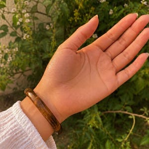 Puede incluir: Una pulsera r&iacute;gida pulida y lisa que presenta un rico patr&oacute;n de piedra ojo de tigre en tonos marr&oacute;n dorado, con bandas naturales tornasoladas en tonos marr&oacute;n claro y oscuro, llevada en la mu&ntilde;eca sobre una prenda blanca texturizada de manga larga.
