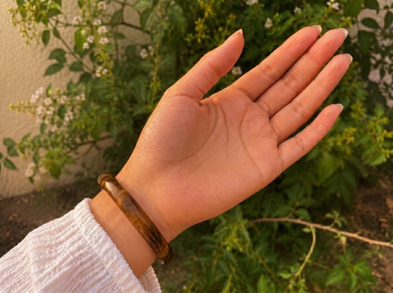 Puede incluir: Una pulsera r&iacute;gida pulida y lisa que presenta un rico patr&oacute;n de piedra ojo de tigre en tonos marr&oacute;n dorado, con bandas naturales tornasoladas en tonos marr&oacute;n claro y oscuro, llevada en la mu&ntilde;eca sobre una prenda blanca texturizada de manga larga.