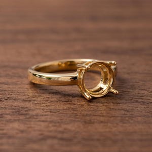 May include: A polished gold ring setting featuring a four-prong head designed to hold a single gemstone. The band is smooth and circular, showcasing a reflective metallic finish throughout the entire structure of the jewelry piece.