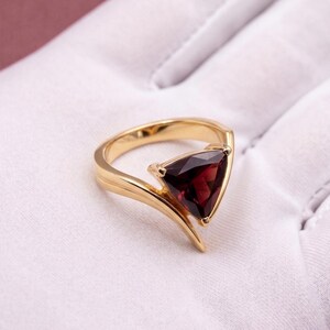 May include: A gold ring featuring a single, deep red, triangular-cut gemstone set in a bypass-style band. The polished gold metal curves around the base of the faceted stone, creating a sleek, open design that highlights the dark crimson color.