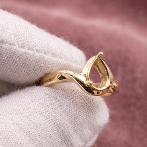 May include: A gold ring setting featuring a bypass-style band that curves upward to form a pear-shaped prong mounting. The polished metal surface reflects light, and the design is intended to hold a single pear-cut gemstone in the center.