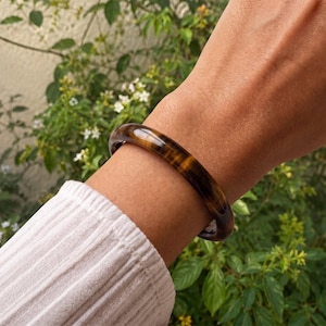 Puede incluir: Un brazalete r&iacute;gido liso y pulido hecho de piedra ojo de tigre, con bandas chatoyantes caracter&iacute;sticas de color marr&oacute;n dorado y marr&oacute;n oscuro que crean una apariencia brillante y estratificada alrededor de la estructura circular s&oacute;lida.