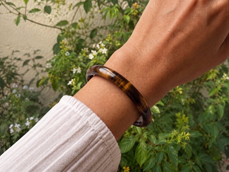 Puede incluir: Un brazalete r&iacute;gido liso y pulido hecho de piedra ojo de tigre, con bandas chatoyantes caracter&iacute;sticas de color marr&oacute;n dorado y marr&oacute;n oscuro que crean una apariencia brillante y estratificada alrededor de la estructura circular s&oacute;lida.