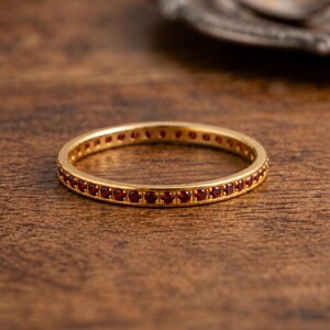 Puede incluir: Un anillo dorado con una banda de pequeñas piedras preciosas rojas. El anillo está sobre una superficie de madera, con un plato plateado borroso en el fondo. Un anillo de eternidad, a menudo para bodas o aniversarios.