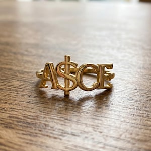 May include: A gold ring spelling "ASCE", with a dollar sign replacing the "S". The ring is displayed on a wooden surface. The gold ring's design is a bold statement piece, perfect for adding a touch of luxury to any outfit.
