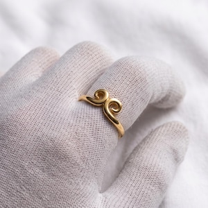 May include: A gold ring featuring a central design of two symmetrical, mirrored swirls that meet at the top of the band. The metal has a smooth, polished finish, and the band is thin and rounded, creating a continuous loop around the finger.