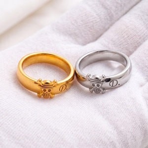 May include: Two rings, one gold-toned and one silver-toned, each featuring a raised skull and crossbones emblem and the letter D followed by a period engraved on the band. The rings are smooth, polished, and have a simple, rounded design.