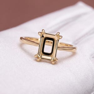 May include: A polished yellow gold ring setting featuring a rectangular head designed to hold a gemstone. The setting includes four prongs at the corners and a smooth, slender band, showcasing a clean, metallic finish without any stone set inside.