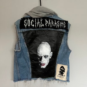 May include: A blue denim vest with a black patch that says "SOCIAL PARASITE" in white lettering. The vest has a black patch with a painted image of a pale-skinned person with a dark background. The vest also has a small white patch with black lettering that says "meth."