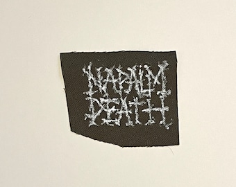 May include: A black fabric patch with the white text "NAPALM DEATH" in a stylized, gothic font. The patch has a slightly irregular shape and is set against a white background.