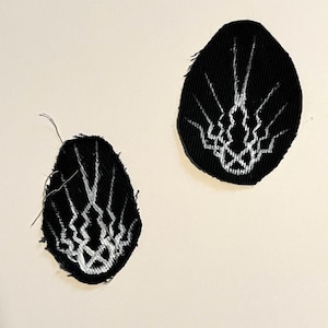 May include: Two black fabric patches with white abstract designs. The patches are oval-shaped and have a textured appearance. The white design features a series of lines and interwoven patterns, creating a unique visual element.