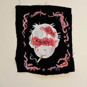 May include: A black fabric patch with a white and red painted image of a person's head with a cigarette in their mouth. The head is surrounded by a pink, swirling design.