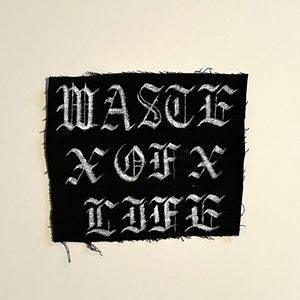 May include: Black fabric patch with white embroidered text that reads "WASTE OF LIFE" in a gothic font.
