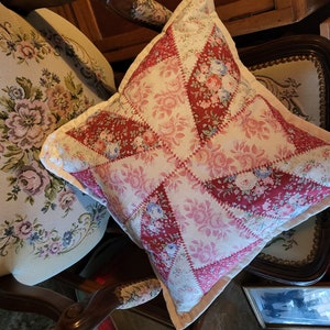 May include: A square cushion with a patchwork design. The cushion features a pattern of red and white fabric squares, each adorned with floral prints in shades of pink, red, and blue. The cushion has a light beige border and is placed on a wooden chair.
