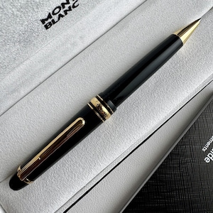 Op de afbeelding: Een zwarte balpen met goudkleurige accenten, voorzien van een goudkleurige clip en een goudkleurige punt. De pen heeft een centrale goudkleurige ring met gegraveerde details. De merknaam Montblanc is zichtbaar in de presentatiebox.