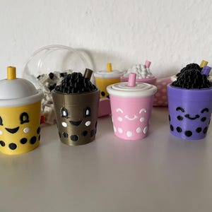 May include: A collection of colorful, smiling boba tea cup figurines. The cups are yellow, brown, pink, and purple, each with a different design. The boba tea toppings are black. Some cups have lids and straws.