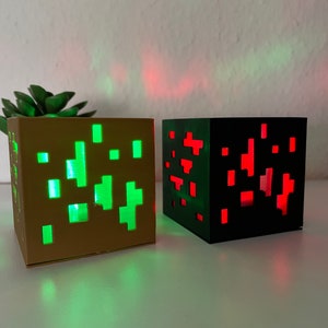 Pode incluir: Dois blocos luminosos com tema Minecraft. O bloco à esquerda é castanho com uma luz verde que brilha através dele. O bloco à direita é preto com uma luz vermelha que brilha através dele.