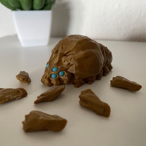 Può includere: Una creatura di colore marrone stampata in 3D con occhi blu e gambe segmentate. La creatura è sdraiata su una superficie bianca con diversi altri pezzi stampati in 3D di colore marrone.