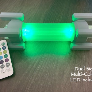 Könnte beinhalten: Eine weiße Plastikleuchte mit einem grünen LED-Licht. Die Leuchte verfügt über eine Fernbedienung mit Tasten für Ein/Aus, Timer, Fade, Jump und Farbauswahl. Der Text "Dual Side Multi-Colour LED included" ist unterhalb der Leuchte aufgedruckt.