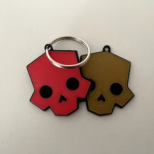 Op de afbeelding: Twee sleutelhangers in de vorm van een schedel, een rood en een bruin, bevestigd aan een zilveren ring.