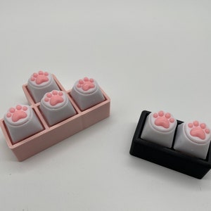 Może przedstawiać: Dwa zestawy klawiszy z różowymi odciskami łapek. Klawisze znajdują się w różowym i czarnym uchwycie. Klawisze są do klawiatury.