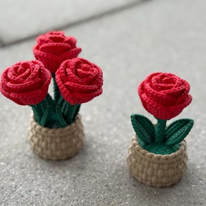 Può includere: Due composizioni di rose rosse all'uncinetto. Una presenta tre rose in un cestino di vimini beige, l'altra una singola rosa. Si vedono steli e foglie verdi.