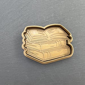 Könnte beinhalten: Ein hölzerner Ausstecher in Form eines Bücherstapels, mit einem offenen Buch oben. Der Ausstecher ist hellbraun und liegt auf einer dunkelgrauen Oberfläche. Das Design zeigt die Seiten und Buchrücken detailliert.