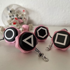 May include: Four pink keycaps with different geometric shapes in white on a black background. The keycaps are attached to strings.