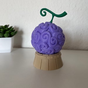 Peut inclure: Une figurine en 3D imprimée en violet et vert d'un fruit avec une tige. Le fruit est sur une base en bois brun.