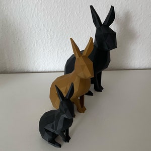 Op de afbeelding: Drie origami-konijnen, één zwart en twee bruin, zijn gerangschikt in een rij van groot naar klein op een wit oppervlak.