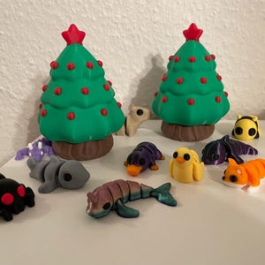 Op de afbeelding: Een verzameling kleurrijke 3D-geprinte figuurtjes, waaronder een zwarte spin, een paars-zwarte dolfijn, een geel kuiken, een paars-zwarte pinguïn, een groen-rode kerstboom, een oranje-witte rups, een paars-zwarte octopus en een wit-bruin hert.