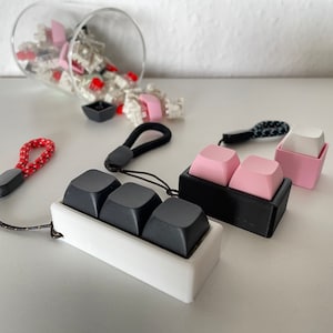 May include: An assortment of keyboard keycaps in various colors, including black, pink, and white. Some keycaps are arranged in small holders, while others are scattered from a tipped-over glass container. A red and black lanyard is also visible.