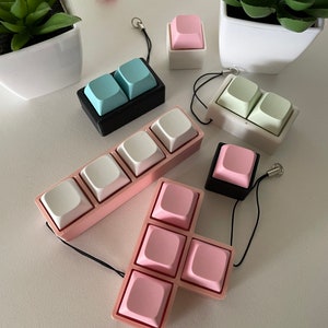 Op de afbeelding: Een verzameling pastelkleurige toetsenbordtoetsaccessoires. De keycaps zijn in verschillende configuraties, waaronder paren en grotere clusters, in tinten roze, wit en blauw. Elke keycap is bevestigd aan een zwart koord.