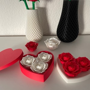 May include: Two heart-shaped boxes with lids, one red and one white, each containing three paper roses. The red box has white roses, and the white box has red roses. There are two additional paper roses, one white and one red, on the table.