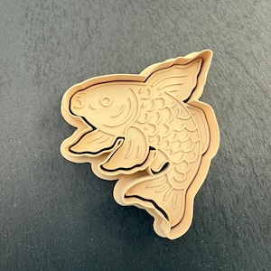 Könnte beinhalten: Ein beiger Kunststoff-Ausstecher in Form eines Goldfisches. Der Ausstecher weist detaillierte Merkmale wie Schuppen, Flossen und Augen auf, mit schwarzen Konturen. Er liegt auf einem dunkelgrauen Hintergrund.