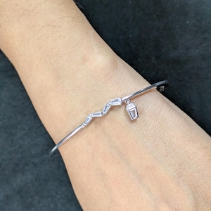 Op de afbeelding: Een zilveren armband met een zigzagontwerp en een kleine bedel. De armband heeft rechthoekige, heldere stenen en een kleine bedel in de vorm van een beker. De armband wordt om de pols gedragen.