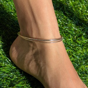 May include: A gold-toned anklet with a delicate design, featuring two thin bands. The anklet is worn on a foot, set against a backdrop of green grass, creating a simple and elegant aesthetic.