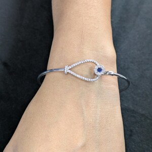 May include: A silver bracelet featuring a teardrop-shaped design at its centre. The teardrop is adorned with small, clear stones and a central blue gemstone. The bracelet is worn on a wrist.
