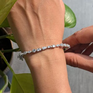 Könnte beinhalten: Ein silbernes Armband mit verschieden geformten, klaren Edelsteinen. Das Armband befindet sich an einem Handgelenk, mit einer Hand, die den Verschluss einstellt. Die Edelsteine umfassen runde, ovale und Baguette-Schliffe. Das Armband ist ein zartes Schmuckstück.