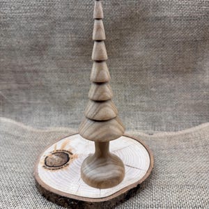 May include: A handcrafted wooden Christmas tree with a tiered design, standing on a round wooden base. The tree is light brown, and the base has a natural wood grain. The tree is approximately 15 cm tall.