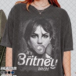 Può includere: T-shirt grigio scuro con un'immagine in bianco e nero di una donna che soffia una bolla. Il testo "It's Britney bitch!" è stampato sotto. Disponibile in vari colori.