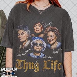 Puede incluir: Camiseta gris oscuro con un gráfico de cuatro mujeres, firmas y el texto "Thug Life" en letras doradas. La camiseta tiene un aspecto vintage y desgastado. Disponible en varios colores.