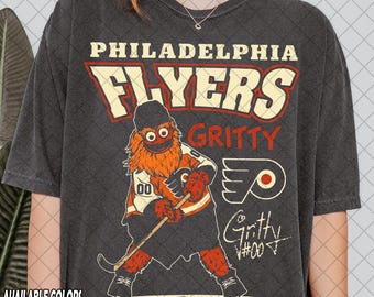 May include: Dark grey t-shirt with the Philadelphia Flyers' mascot, Gritty, in hockey attire. The design includes the team name, mascot's name, and logo. Available in various colours, this shirt is a fan item.