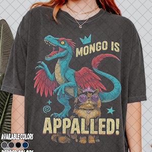 May include: Dark gray t-shirt featuring a cartoon dinosaur with blue and red accents and a cat wearing sunglasses and a tiara. Text reads "MONGO IS APPALLED!" Available in several colors.
