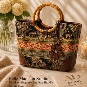 May include: A small tote bag featuring a patterned fabric with monkeys and trees, brown leather trim, and two circular bamboo handles. A brown leather flower charm hangs from one handle. The bag displays the text Relic Madison Studio Monkey Elegance Bamboo Handle Small Tote.