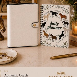 Könnte beinhalten: Ein weißes Coach-Notizbuch mit schwarzem Rand und Druckknopfverschluss, kombiniert mit einem spiralgebundenen Tagesplaner mit Pferdemuster und dem Text daily planner. Ein goldener Coach-Stift liegt daneben, zusammen mit einer Schale mit Pferdeanhängern.