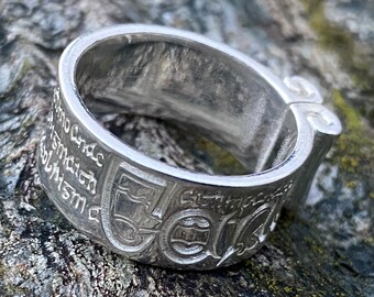 May include: A silver ring with an inscription around the band. The inscription is in a foreign language. The ring is open on one side and has a hinge.