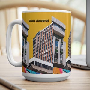 May include: White ceramic mug with a design of a building and the text "Glasgow, Strathclyde Uni" on a yellow background. The mug is on a wooden surface with a pen and a laptop in the background.