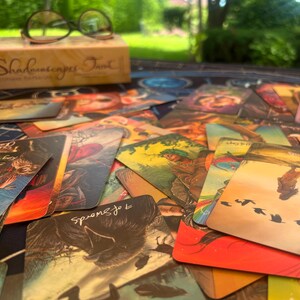 May include: A collection of Shadowscapes Tarot cards spread out on a surface, featuring colorful illustrations of figures, animals, and nature scenes. A box labeled Shadowscapes Tarot and a pair of black-rimmed glasses sit in the background.